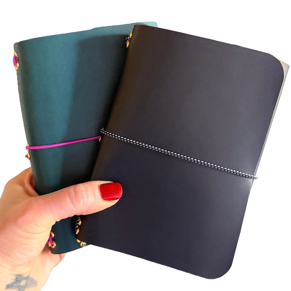 Two hardcover notebooks held in a hand against a white background. The front notebook is dark charcoal gray with a silver elastic closure, and the back notebook is teal with a pink elastic closure and gold-toned embellishments.
